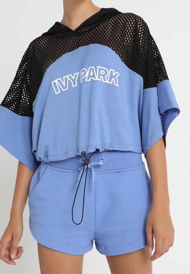 Blå Ivy Park-shorts och matchande kort topp med svart mesh-hood, bärs av en person med avslappnade händer vid sidorna.
