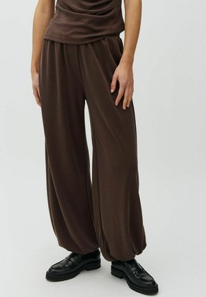 Personne portant un pantalon marron ample avec des poignets froncés et des mocassins noirs à semelle épaisse, debout sur un fond uni.