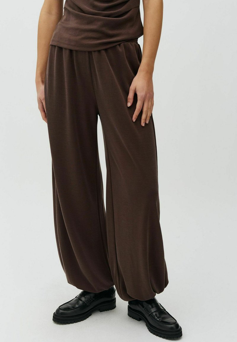 Personne portant un pantalon marron ample avec des poignets froncés et des mocassins noirs à semelle épaisse, debout sur un fond uni.