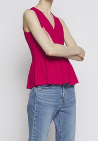 Personne portant un top peplum rouge sans manches et un jean bleu, bras croisés, sur un fond gris clair uni.