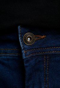 Jeans de mezclilla azul con un botón de metal de tono cobre que presenta grabado "TOM TAILOR". Costuras visibles en un color amarillo-marrón contrastante.