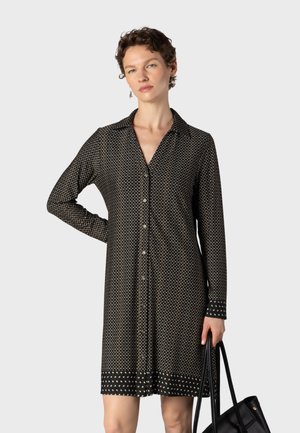 Femme aux cheveux courts et bouclés portant une robe noire à motifs, manches longues et boutonnée, tenant un sac fourre-tout noir sur un fond uni.
