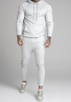 Mand iført tætsiddende helt hvid hættetrøje og joggingbukser med tatoveringer på hænder og hals, stående mod en ensfarvet grå baggrund.