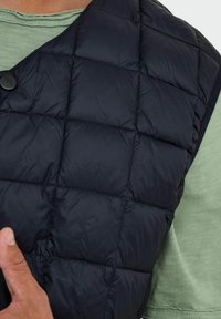 Gilet matelassé bleu marine avec une texture douce, présentant de grands motifs de couture et une fermeture à boutons, porté sur une chemise vert clair.