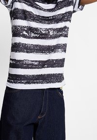 Pruhovaný tričko s černými a bílými horizontálními štětcovými vzory. Krátké rukávy a volný střih, spárované s tmavými džínami.