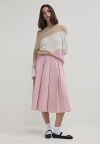 Donna che indossa un maglione a blocchi di colore, una gonna midi plissettata rosa, calze bianche e ballerine nere in stile Mary Jane, in piedi contro uno sfondo bianco.