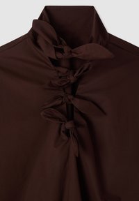 Camicia marrone con collo alto e laccetti annodati sul davanti. Tessuto liscio e strutturato. Design semplice senza motivi o decorazioni.