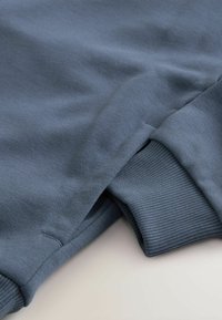 Sweatshirt bleu au toucher lisse, avec des poignets côtelés et une coupe décontractée. Le matériau semble doux et légèrement extensible.