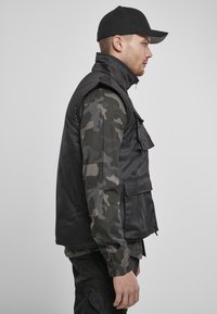 Svart polstret vest med høy krage, sidelommer og glatt overflate, brukt over en grå camo-langermet skjorte, vist i profil.