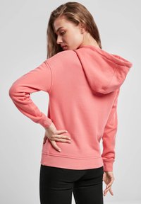 Felpa rosa con cappuccio regolabile, polsini e fondo a costine. Tessuto liscio; indossata con pantaloni neri, mostrando una vista laterale e posteriore.