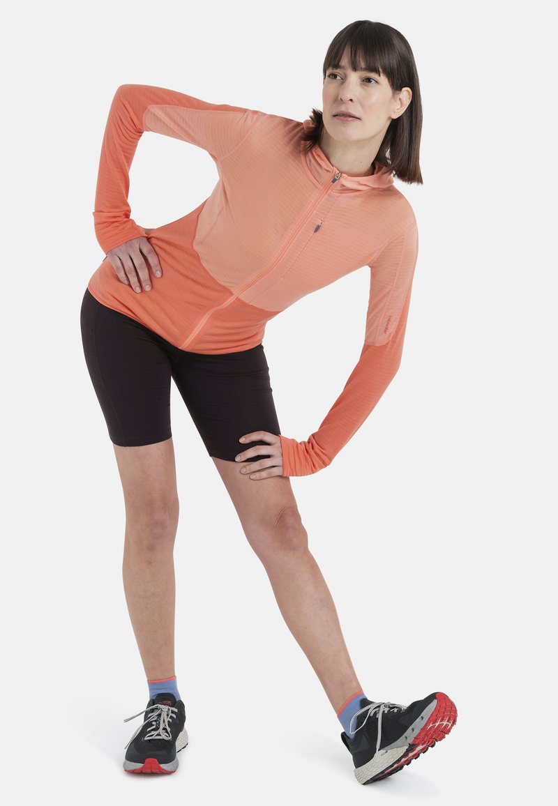 Veste légère à manches longues zippée de couleur orange vif avec un motif texturé, associée à un short noir et des chaussures de course noires avec des accents rouges.