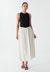 Mustat hihattomat topit, joissa on tekstuuri, yhdistetty virtaavaan off-white midi-hameeseen. Mallilla mustat remmisandaalit.