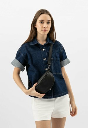 Denim overhemd met knopen en opgerolde mouwen, gecombineerd met witte shorts. Een kleine, zwarte leren schoudertas wordt met één hand vastgehouden.
