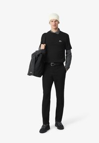 Camiseta negra de manga corta, camiseta gris en capas por debajo, pantalones negros, gorro blanco, llevando una chaqueta gris y una bolsa negra, zapatillas negras.