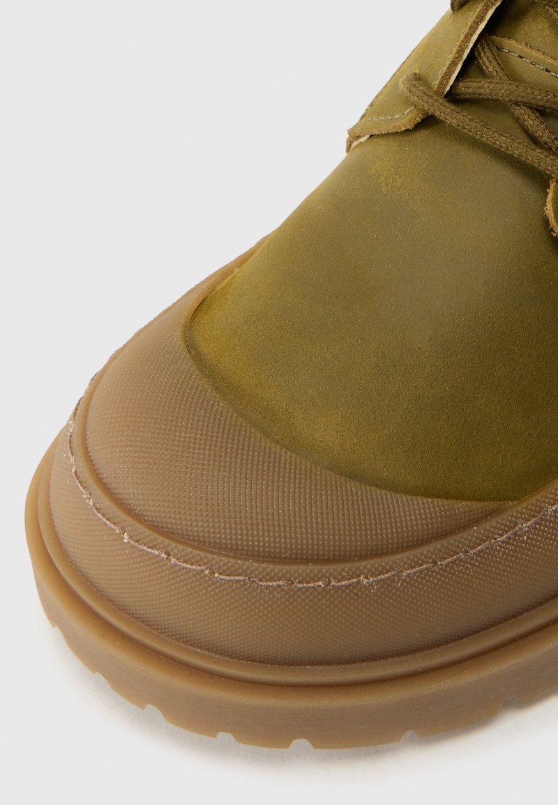 Zapato de cuero verde con una suela de goma texturizada y marrón claro. Punta redondeada, costuras en contraste y acentos sutiles en el material superior.