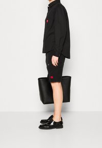 Man som står på sidan iförd svart långärmad skjorta, svarta cargoshorts, svarta blanka skor, och håller en stor svart tygkasse framför en vit bakgrund.