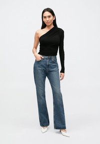 Čierny top na jedno rameno s nariasenou textúrou, skombinovaný s vysokým pásom, džínsami s rovnými nohavicami modrej farby a bielymi lodičkami so špicatou špičkou.