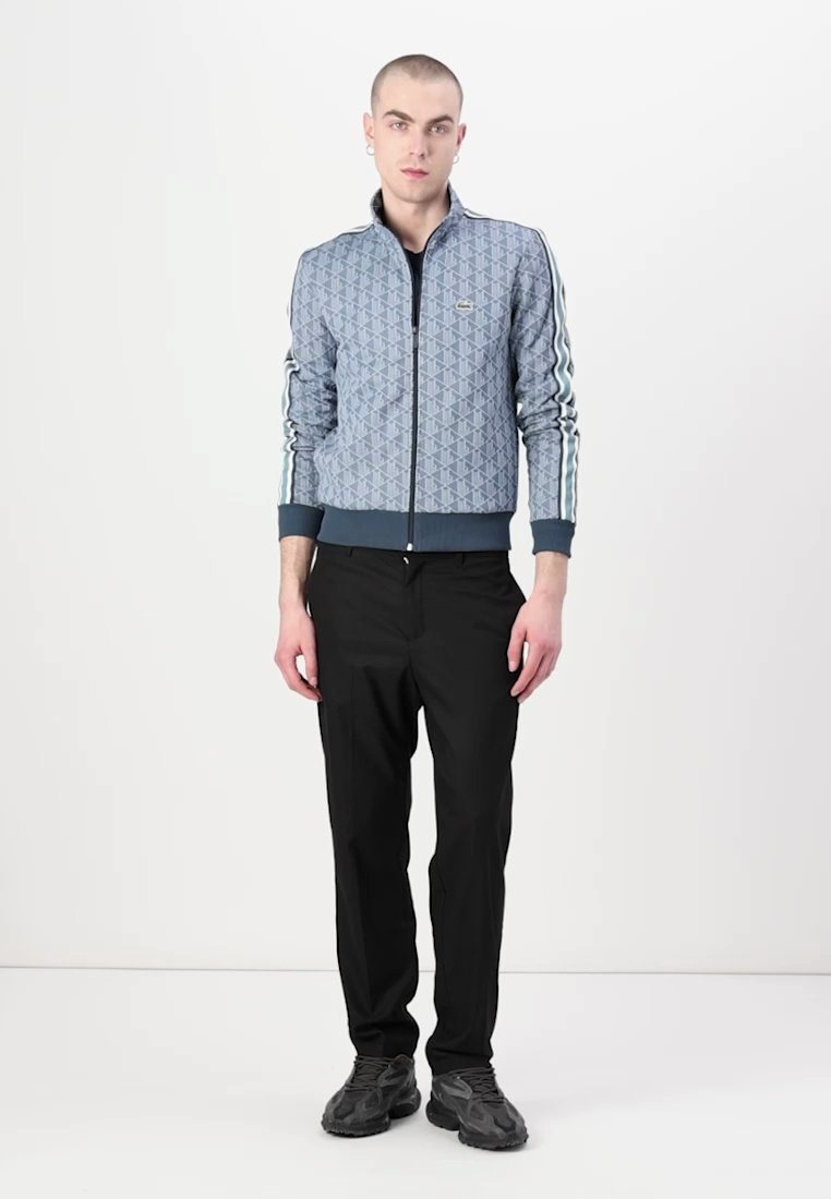 Veste bleue à motif en tissu texturé, avec fermeture éclair à l'avant et accents rayés, associée à un pantalon noir et des baskets sombres.