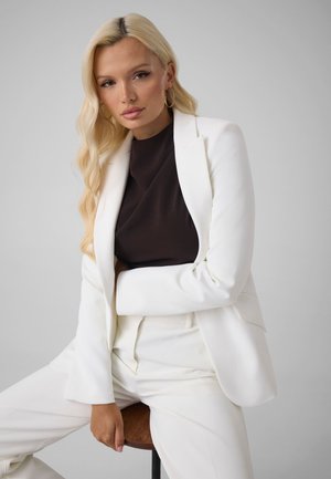 Mujer rubia con cabello largo y ondulado, vestida con traje blanco y top negro, sentada en un taburete de madera contra un fondo liso.