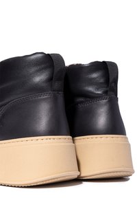 Par de botas de tobillo de cuero negro con suelas de goma gruesa color marrón y tiradores de tela negra en la parte trasera, mostradas desde un ángulo trasero.