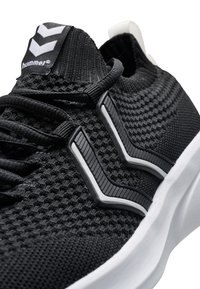 Svart idrottssko med texturerad stickad ovansida, vit sula och framträdande grå accenter i ett zick-zackmönster. Har platta skosnören och logotags.