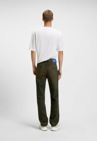 Camiseta blanca de manga corta combinada con unos jeans verde oscuro, ligeramente desgastados, que cuentan con una etiqueta azul en la cinturilla y dos bolsillos traseros.