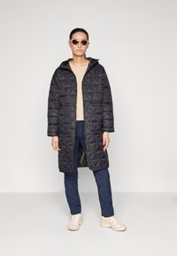 Mustat tikatut pitkät takki hupparilla, joissa on vetoketjullinen etuosa ja taskut, puettuna beige paidan ja tummansiniset housujen yli, yhdistettynä aurinkolaseihin.