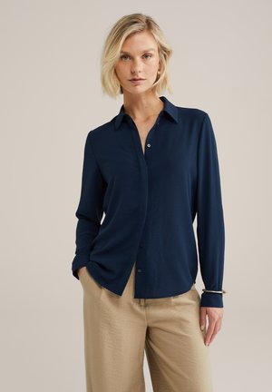 Blonde vrouw met kort haar, gekleed in een marineblauw knoopblouse en beige broek, staande met één hand in de zak tegen een beige achtergrond.