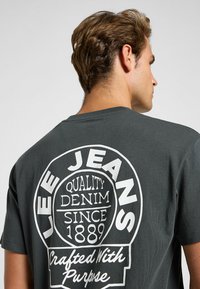Camiseta de algodón gris oscuro con un gran logo impreso en blanco en la parte posterior que dice: "LEE JEANS, QUALITY DENIM, SINCE 1889, Crafted With Purpose."