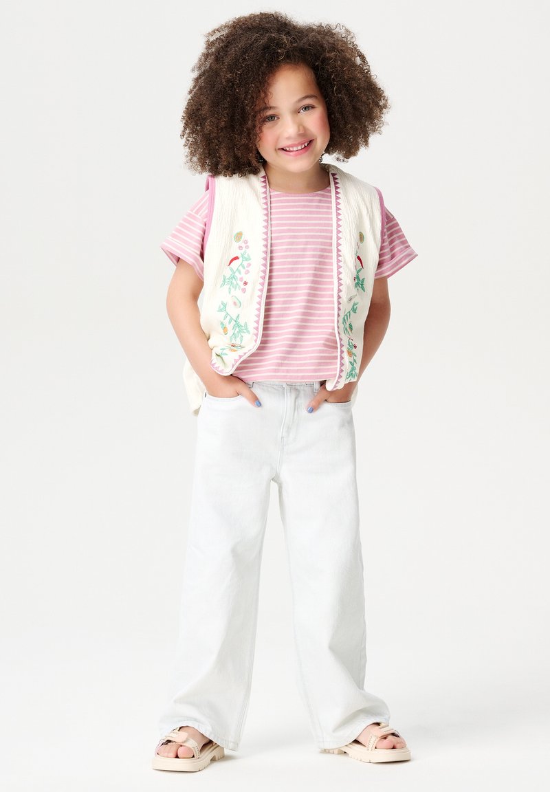 Witte geborduurde vest over een roze gestreept T-shirt, lichtblauwe wijde spijkerbroek en beige sandalen met een gesp. Korte mouwen en een relaxte pasvorm.