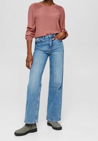 Staubrosa Bluse mit langen Ärmeln und plissierter Textur an den Schultern, kombiniert mit hellblauen weitgeschnittenen Jeans und grauen Stiefeletten.