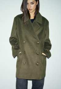 Olive zeleno dvoredno volneno plašč s velikimi gumbi, stranskimi žepi in širokim ovratnikom. Ima sproščen kroj in gladko teksturo.