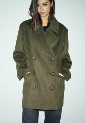 Cappotto di lana doppiopetto verde oliva con grandi bottoni, tasche laterali e un ampio colletto. Presenta una vestibilità comoda e una consistenza liscia.