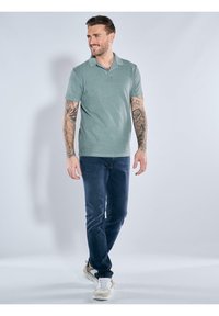 Green short-sleeve polo shirt with collar, paired with dark jeans and beige athletic shoes. Fabric appears smooth with a consistent texture.