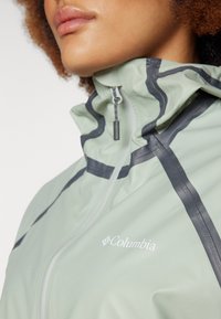 Veste imperméable vert clair avec un col haut, des rayures noires contrastantes et une fermeture éclair ; présente un logo sur la zone gauche de la poitrine.
