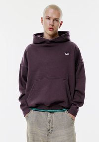Haut à capuche oversized violet avec une poche avant et un petit logo sur la poitrine, associé à un pantalon gris clair avec une taille rayée.