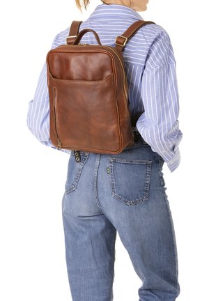 Donna che indossa una camicia a righe blu e jeans con uno zaino in pelle marrone dotato di tasca frontale con cerniera e spallacci regolabili.