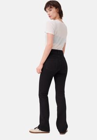 Donna dai capelli corti castani che indossa una maglietta bianca trasparente, jeans neri a vita alta e sneakers bianche, in piedi e guarda oltre la spalla.