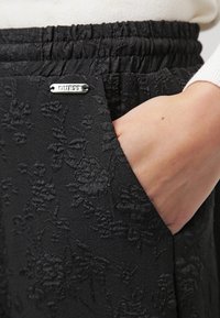 Zwarte stoffen broek met een reliëf bloemenpatroon, elastische tailleband en een zilveren logo-accent nabij de zak.
