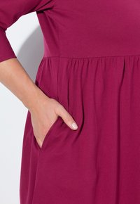 Fuchsiové šaty s tříčtvrtečními rukávy, nařaseným pasem a bočními kapsami. Vyrobeno z měkké látky s hladkým povrchem.