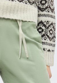 Πλεκτό πουλόβερ με ασπρόμαυρο σχέδιο συνδυασμένο με ανοιχτοπράσινο παντελόνι με κορδόνια που γράφουν "Marco Polo" και "Γεια, με λένε Marithe."