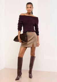 Olkapäät paljaat tummanvioletti kaapelineulepaita, ruudullinen minihame, jossa edessä halkio, ruskeat nahkaiset polven yli ulottuvat saappaat, ja ruskea pikkulaukku.