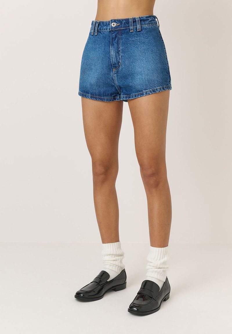 Șorturi din denim cu talie înaltă și margine destrămată, având o culoare albastră clasică și un design cu cinci buzunare. Purtați cu mocasini negri din piele lucioasă și șosete albe.