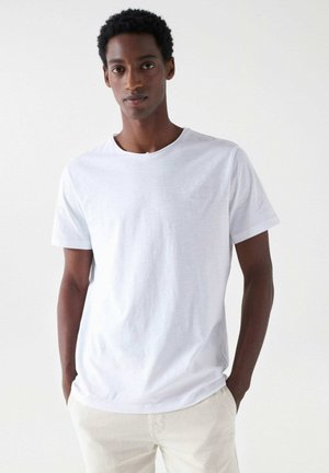 Camiseta de algodón blanca de manga corta, con cuello redondo y un sutil detalle de logo en el pecho. Textura suave y corte relajado.