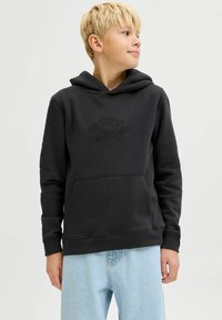 Melnā kapucēta džaka no mīkstas auduma, ar kangaru kabatu, ribinātiem piedurknēm un reljefā "ORIGINALS" logotipā uz priekšpuses, kombinēta ar gaiši zilām džinsām.