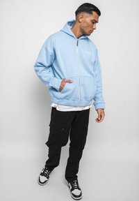 Light blue zip-up hoodie with a front pocket and embroidered logo, paired with black cargo pants and black sneakers with white accents.