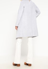 Cappotto grigio chiaro realizzato in tessuto liscio, con maniche lunghe e una silhouette leggermente svasata; indossato su pantaloni bianchi svasati con tacchi marroni.