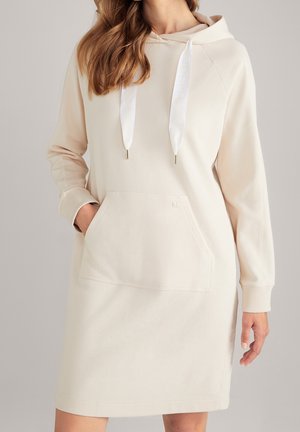 Kobieta ubrana w kremową sukienkę-bluzę z białymi sznurkami i przednią kieszenią kangurką, stojąca z jedną ręką w kieszeni.