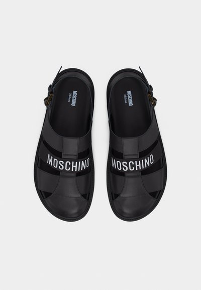 Paire de sandales en cuir noir avec brides de cheville ajustables et logo "MOSCHINO" sur les brides avant, vues de dessus.