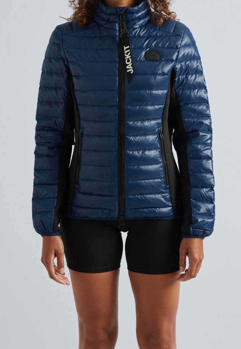 Femme portant une veste matelassée bleue avec fermeture éclair et panneaux latéraux noirs ainsi qu'un short noir, debout devant un fond gris uni.